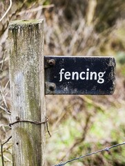 Weathered Wooden Post Supporting a Rustic Metal Fencing Sign in Natural Surroundings with Blurred Background and Greenery in Rural Environment