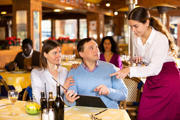 Positive couple visiting cozy restaurant for lunch, consulting with hospitable young waitress while making order..