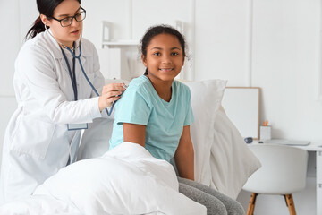 Fototapeta premium Female doctor with stethoscope listening to African-American girl in hospital room