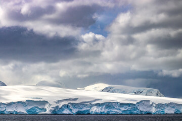 Shoreline of the Antarctic peninsula covered in deep snow. Ocean in foreground; blue sky and clouds behind. 
