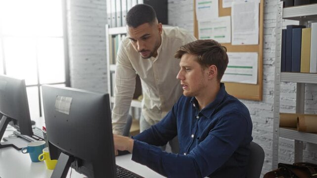 Men working together in a modern office, discussing tasks on a computer screen, displaying a collaborative business environment with paperwork on the walls and shelves of files in the background.