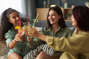 Cheerful young ethnically diverse gen Z girls sitting on couch in living room, clinking glasses with soft drinks during party