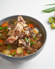 A bowl of savory meat and vegetable soup showcases tender chunks of meat, carrots, potatoes, and vibrant green herbs, accompanied by fresh citrus