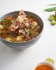 A hearty bowl of beef soup filled with tender meat, potatoes, carrots, and topped with fresh herbs, served with lime and spicy sauce