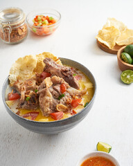 A vibrant bowl of savory soup with tender meat, fresh tomatoes, herbs, crispy chips, refreshing limes, and tangy dip