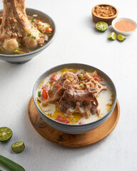 A bowl features rich soup with tender meat, colorful vegetables, fresh lime halved, and sauces served on a wooden surface