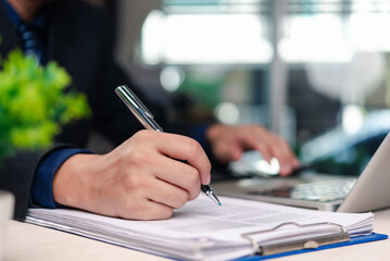 Businessman signing documents working on laptop in office environment. Close-up view of executive hand writing with pen on paper documents beside computer workspace.