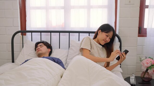 Asian woman secretly chatting to lover while lying in bed with husband. 