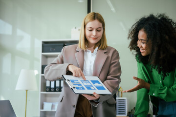An African American businesswoman and a Caucasian woman collaborate at a desk, brainstorming...