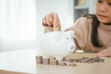 A girl sits at a table, saving money by inserting coins into a piggy bank, emphasizing the importance of building a strong financial foundation for a secure future