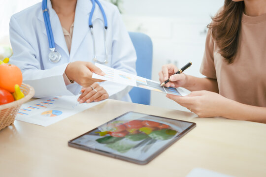 A young female nutritionist expert talks to a female patient at a table, recommending a healthy diet with vegetables and fruits to promote wellness, balanced nutrition, and effective weight loss
