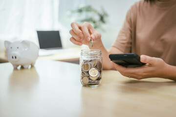 A young woman saves money for the future by putting coins in a piggy bank on the table, managing monthly expenses, and focusing on investment and business growth for financial stability