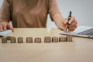 A young woman saves money for the future by putting coins in a piggy bank on the table, managing monthly expenses, and focusing on investment and business growth for financial stability