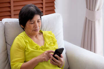 Senior asian woman sitting on sofa looking smartphone for video call in living room at home, elderly using social media on smart phone for chat online and conversation distant, lifestyle concept.