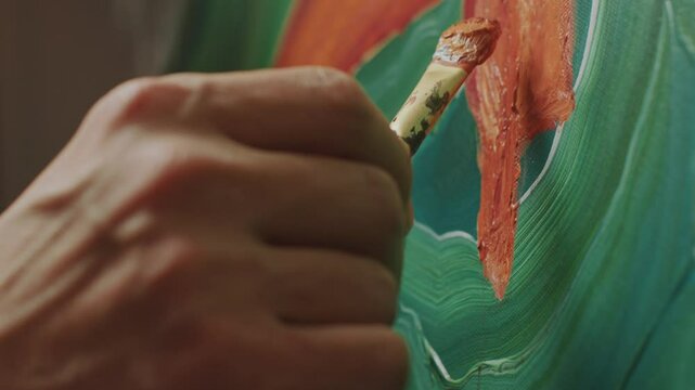 Hands of unrecognizable male artist applying orange paint to canvas with brush and holding palette of mixed colors, working on new painting in studio. Close-up shot