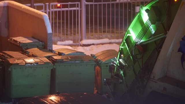 Street light illuminating green garbage bins in snow. Removing municipal solid waste from residential collection site with garbage truck.