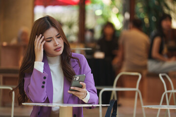 Young asian businesswoman looking worried while reading a message on her phone, sitting at a table in a cafe, touching her head with one hand