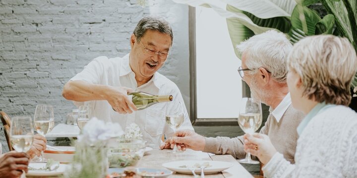 Asian man pouring wine for an retired couple. Man pouring wine is smiling. The retired couple enjoys wine at a table. Wine and dining scene with friends. Diverse retired friends having wine for lunch.