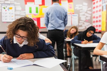 Students in a classroom focused on writing. Diverse group, including a girl in a hijab. Classroom setting, students writing, engaged in learning. Girl in exam at school.