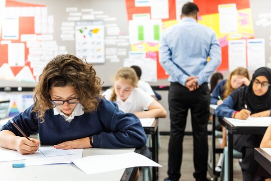 Focused students in a classroom writing and taking an exam. Diverse students in uniforms taking exam. Classroom exam setting with attentive students and teacher. Teacher look out students taking exam
