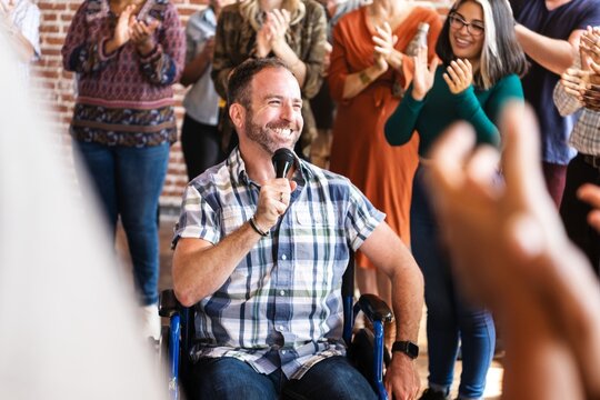 Man in wheelchair speaking to microphone in public with microphone, surrounded by clapping crowd. Joyful and supportive atmosphere, engaging speaker. Inclusive, diverse audience, lively event.