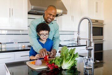 Happy man helps child chop vegetables in a kitchen. Father and son preparing food together on the counter. Cooking, learning, bonding moment. African American father teach his son cooking in kitchen