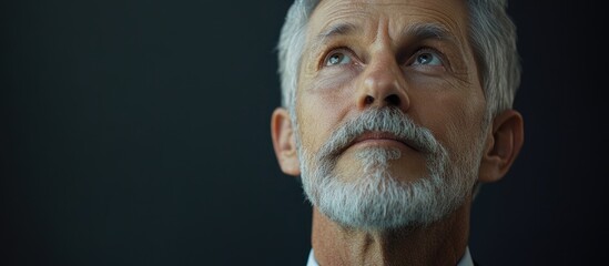 Obraz premium Elderly man with grey hair and beard in a suit looking up against dark background conveying wisdom and experience with calm expression