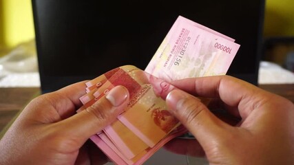Asian man counting income of hundred thousand rupiah Indonesian currency in front of laptop