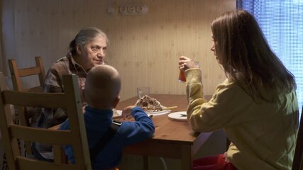 Older woman grandmother sits at table with her young grandson and young woman. Family having evening meal with dessert with light from overhead light . Room simply decorated, but warm and welcoming.