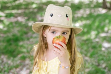 Cheerful little girl in a straw hat playfully holding a bright red apple in a sunny blooming garden.