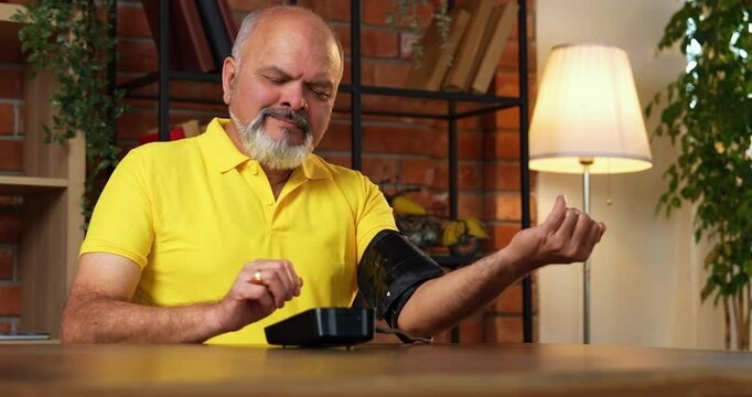 Indian senior man checks blood pressure at home with a digital monitor, sees normal reading, and smiles while holding a pill bottle for routine health care