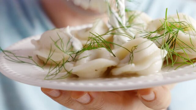 man with Fork takes and eats dumplings from the plate. Boiled dumplings with dill and cream are arranged in a circle on a white plate.