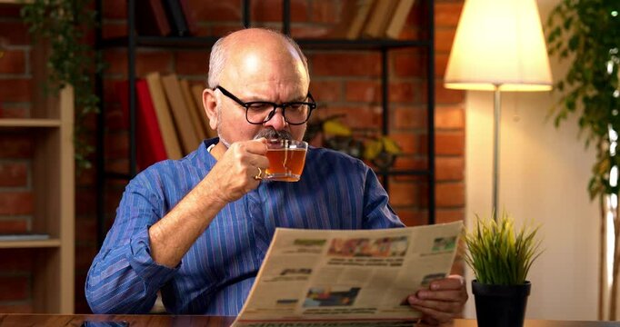 Indian senior retired man wears kurta, reads morning newspaper while sipping black coffee at home desk, staying updated on current business affairs and feeling relaxed