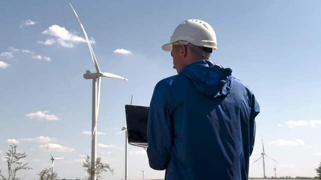 Worker inspects windmill renewable energy site. Wind industry professional monitors turbine performance laptop. Windmill worker ensures operational efficiency. Sustainable wind industry worker field.