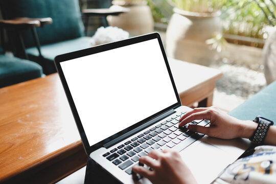 A person typing on a laptop with a blank screen in a cozy seating area featuring wooden tables and soft green furniture, creating a comfortable working environment. - Powered by Adobe
