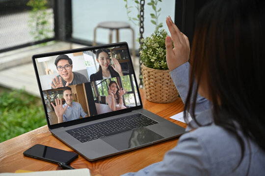 Virtual Team Meeting in a Digital Workplace, A woman in a striped shirt participates in a video conference, waving at four colleagues displayed on her laptop screen