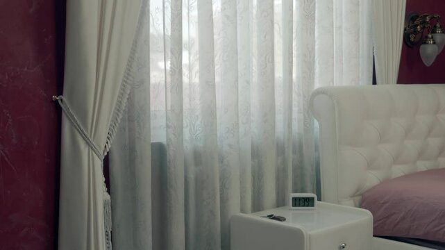 In bedroom white nightstand with a clock, window with beautiful white tulle and curtains hanging on a golden cornice and a white glossy stretch ceiling. Shot in motion