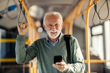 Portrait of an old commuter riding public bus and using his cellphone while smiling at camera.