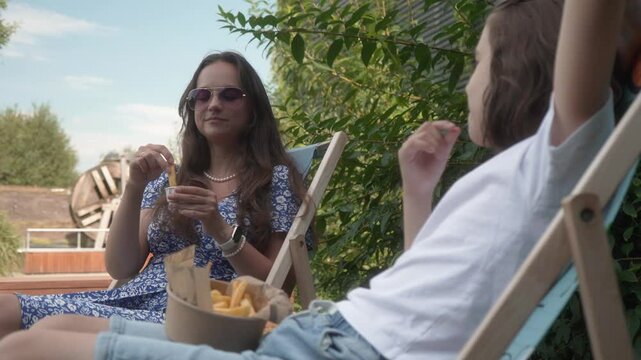 A woman and a child are sitting in lounge chairs in a public park, eating french fries from a disposable paper takeout container. The mother and son are having a fast food snack outdoors.