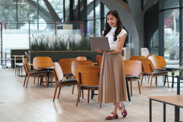 Young woman standing in a contemporary cafe, using a laptop. The setting features stylish wooden chairs and tables, creating a productive and relaxed atmosphere for work