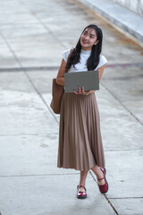 Fototapeta premium Asian businesswoman standing outdoors in elegant casual wear with pleated skirt and comfortable shoes. Working on laptop while carrying tote bag. Modern lifestyle concept of remote work and mobility