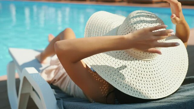 Woman lounging on poolside chair wearing wide-brimmed sun hat and shielding face from sun. Blue water reflects bright sunlight. Concept of summer, vacation, and relaxation