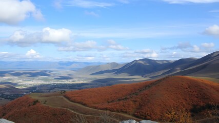 Panoramic view of autumnal hillside overlooking a valley town nestled amongst majestic mountains under a vibrant blue sky.