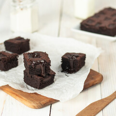 Three rich chocolate brownies stacked on a wooden board, adorned with melting chocolate, accompanied by glasses of milk