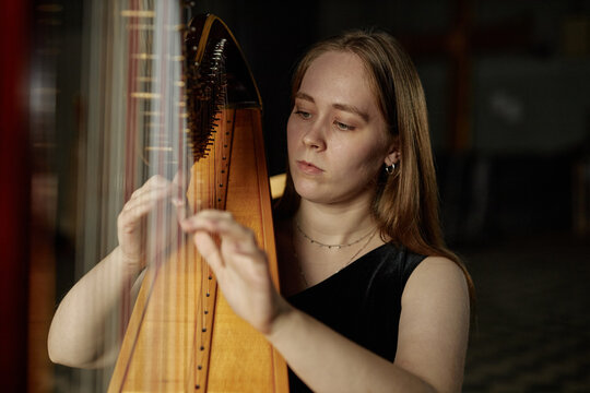 Portrait of professional female harpist performing classical music on string instrument in dimly lit church with excellent acoustics