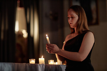 Medium shot of young woman in elegant black dress putting out candle while praying in church, copy space
