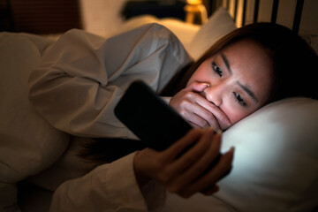 Young Asian female lying under blanket on bed and watching horror movie on mobile phone with a worried expression, illuminated by the screen's glow in a dimly lit room. Dark bedroom in late evening