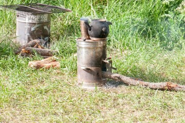 A makeshift outdoor stove heats a kettle over a small fire, nestled in a grassy field. Simple cooking setup.