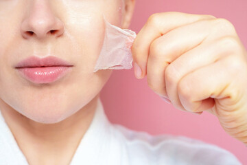 The woman tears off pore cleansing mask from her face on pink background close-up.