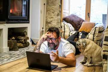 The man is lying on the floor in his living room, talking to his dog, who listens tenderly as they share a peaceful moment. He browses the web on his laptop while a fire burns in the cozy space